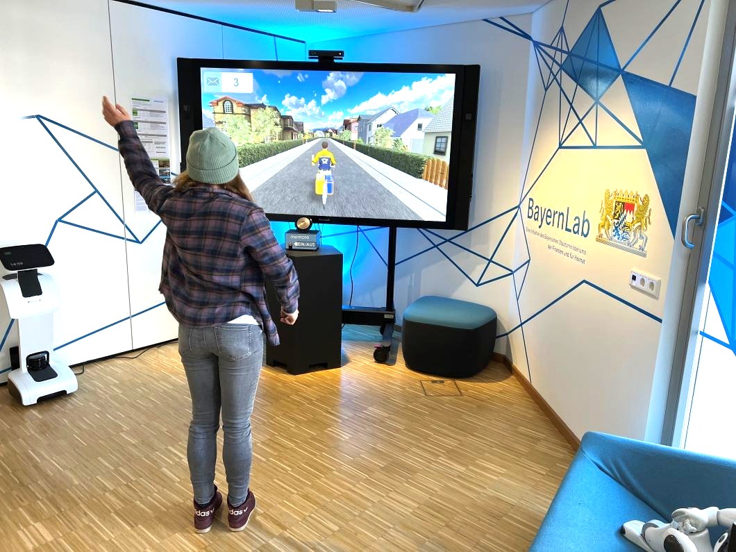 Eine Frau mit Mütze steht vor einem großen Bildschirm und hebt die linke Hand. Auf dem Bildschirm fährt ein Postbote mit dem Fahrrad durch eine Straße und hebt auch die linke Hand